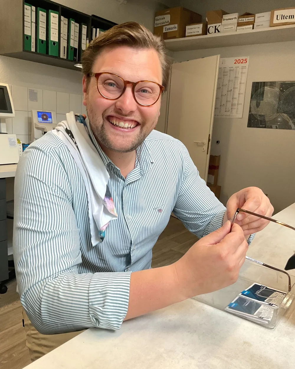 Ein Augenoptiker mit Hornbrille sitzt in der Werkstatt am Tisch und arbeitet an einer Fassung