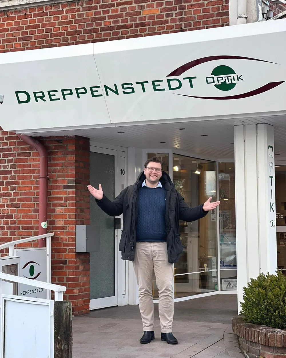 Ein Mann breitet vor dem Eingang von Dreppenstedt Optik einladend die Arme aus, darüber der Schriftzug des Ladens
