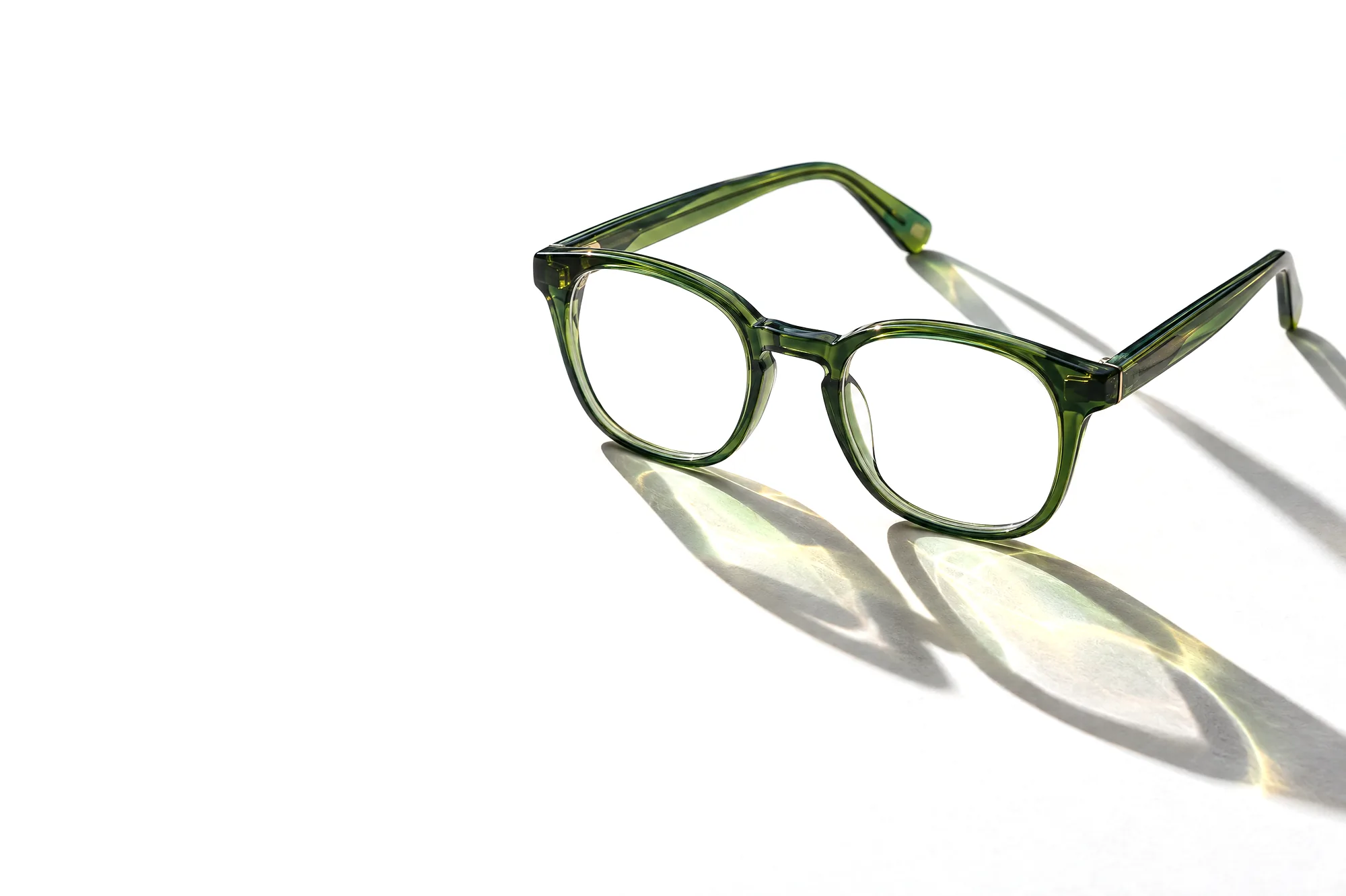 Grüne Acetatbrille schräg im Gegenlicht, ihre Schatten zeigen grüne und goldene Lichtflecken auf hellem Papier
