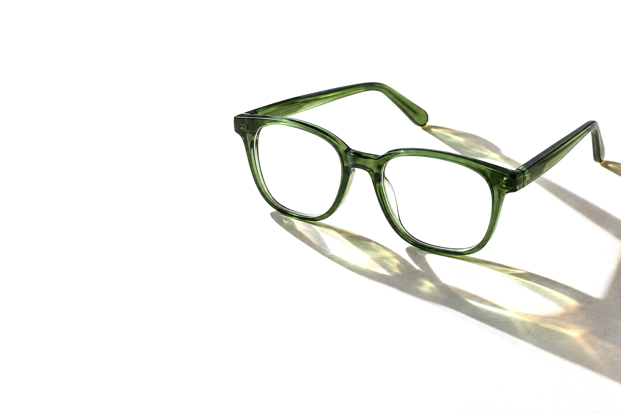 Grüne Acetatbrille im Gegenlicht, ihr Schatten wirft grüne und goldene Lichtflecken auf helles Papier