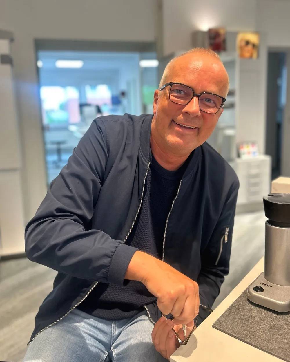 Ein Augenoptiker mit Brille sitzt lächelnd am Arbeitsplatz im Laden, eine Brille in der Hand